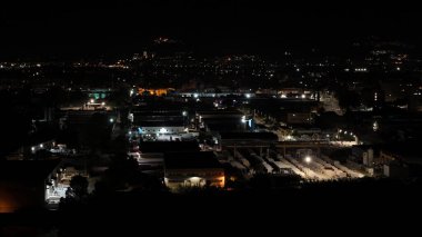 Gecenin örtüsü altında, endüstriyel bölge dağınık ışıklarla parıldıyor, uzak tepelerin arka planına karşı hareketli bir duvar halısı ortaya çıkıyor..