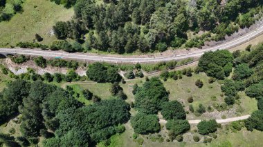 Geniş bir manzara, altın öğleden sonra güneş ışığıyla aydınlatılan yemyeşil ormanlarda kendi yolunu takip eden bir demiryolu hattını ortaya çıkarıyor..