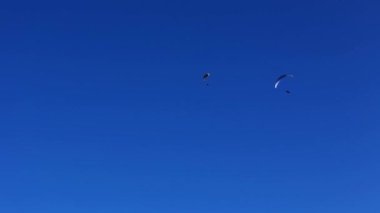 Paraglider mavi gökyüzünde akrobatik bir gösteri sergiliyor. Bu arada başka bir paraglider uzaklarda uçuyor.