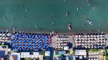 İnsanlar yüzüyor, güneşleniyor ve kalabalık bir plajda mavi şemsiyelerle eğleniyorlar.