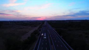 Arabalar ve kamyonlar gün batımında otoyolda şehre doğru ilerliyorlar.