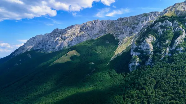 Güneş ışığı görkemli bir dağ sırasını aydınlatıyor. Aşağıdaki yemyeşil ormana gölgeler düşürüyor.