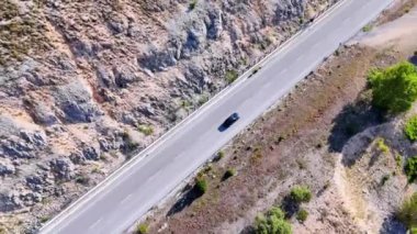 Siyah araba, dağlık bir arazinin içinden geçen bir otoyol boyunca ilerliyor.