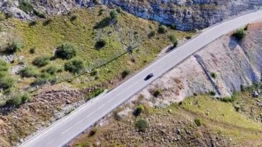 Siyah araba, dağlık bir arazinin içinden geçen bir otoyol boyunca ilerliyor.
