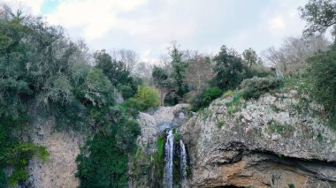 Canlı yeşillikler ve sessiz toprak tonlarıyla çevrili engebeli bir uçurumdan aşağı nefes kesici bir şelale düşer. Sahne, suyun doğal bir havuza nazikçe akması gibi doğanın huzur dolu özünü yakalar.