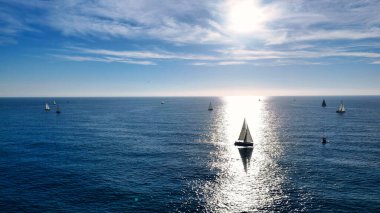 Yelkenliler sakin sularda yüzerler. Beyaz yelkenleri okyanuslardan yansıyan altın güneş ışığıyla aydınlanır. Ufuk mavi tonlarda boyanmış, kabarık bulutlar dağılmış.