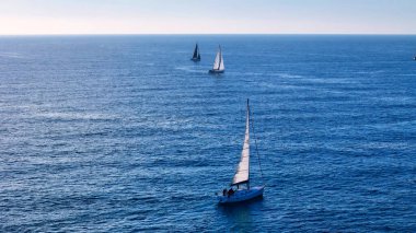 Üç yelkenli zarif bir şekilde masmavi sularda yüzüyor, beyaz yelkenleri hafif rüzgarda dalgalanıyor. Güneş sıcacık bir ışıltı saçar, parıldayan denizi aydınlatır ve sonsuz bir şekilde uzanır.