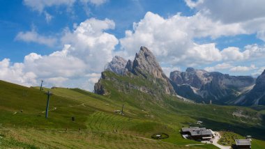 Dolomite dağlarının parlak mavi gökyüzü altındaki nefes kesici manzarası sahneyi dolduruyor. Yumuşak yamaçlar ve yeşil otlar bu huzurlu ortamda gezginler ve kaşifler için davet edici bir alan yaratır..