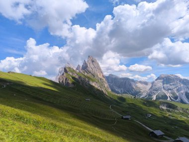 Yüksek kaya oluşumları, parlak güneş ışığının kabarık bulutlara karşı zengin zıtlıklar yarattığı Dolomitler 'deki verimli, yuvarlanan tepelerin üzerinde yükselir. Huzurlu bir atmosfer keşfi ve merakı davet eder.