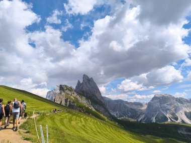 Dolomitler 'in dolambaçlı yollarında gezen maceraperestler, yemyeşil tepeler ve bulutlarla benekli parlak mavi gökyüzünün altında yükselen zirvelerle çevrili..