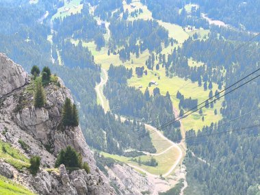 Kayalık bir dağın zirvesinden nefes kesici bir vadiye tepeden bakan yeşil tarlalar aşağıya doğru uzanıyor, uzun çam ormanları boyunca kıvrılan patikalar berrak mavi gökyüzünün altında..