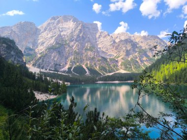 Kristal berraklığında bir göl üzerinde nefes kesici dağlar kulesi, canlı yeşillik sahne çerçevesi ile. Yumuşak bulutlar canlı, mavi bir gökyüzünde sürükleniyor. Gün ortasında huzurlu bir atmosfer yaratıyor..