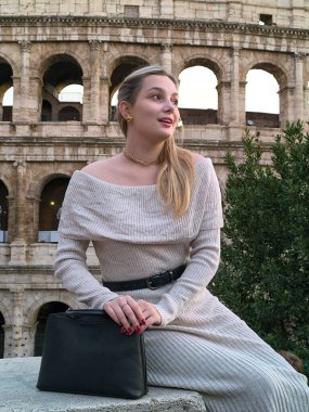 Akşam güneşinin yumuşak parıltısı altında şık bir kadın zarif bir şekilde Colosseum 'un yanında oturuyor. Zarif kıyafetleri tarihi arkaplanı tamamlıyor ve çarpıcı bir görsel ahenk oluşturuyor..