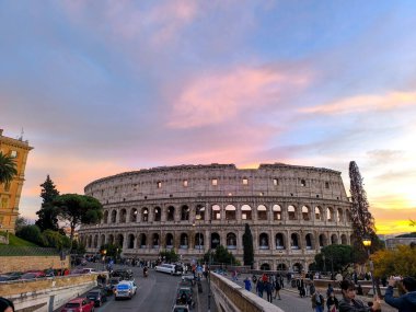 Güneş Roma 'nın üzerinde batarken Kolezyum görkemli bir şekilde duruyor. Etrafı turistler ve tarihi caddelerle çevrili. Gökyüzü çarpıcı bir renk paleti sergiliyor, atmosferi yükseltiyor..