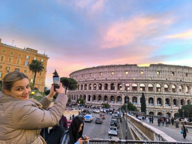 Bir gezgin, gün batımının sıcak gölgesinde yıkanmış çarpıcı Colosseum 'un fotoğrafını çekerken gülümsüyor. Titreyen gökyüzü büyüleyici atmosfere ekleniyor..