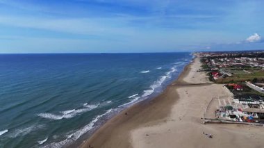 Birkaç kişi kıyı şeridi boyunca yürürken, yumuşak dalgalar altın kıyıya çarpıyor. Parlak güneşin altında parlak bir ufuk uzanıyor..