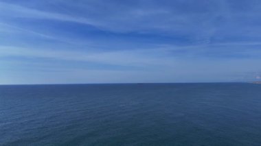 Geniş bir kıyı şeridine vuran hafif dalgalar, güneşin sıcacık parıltısını yansıtıyor. Issız bir gemi uzaklarda görülebilir, bu huzurlu günde açık denizin dingin güzelliğini arttırır..