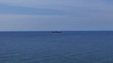 Büyük bir kargo gemisi, sakin mavi okyanusta seyrediyor. Dalgalar gövdesine çarpıyor. Gökyüzü aydınlık ve berrak, huzur ve sükunet hissi veriyor..