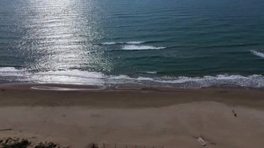 Gün ortası güneşinin altında, yumuşak dalgaların kumlu sahili öptüğü sakin bir sahil uzanır. Işıldayan yüzey ışığı yansıtıyor, barışçıl bir atmosfer yaratıyor..
