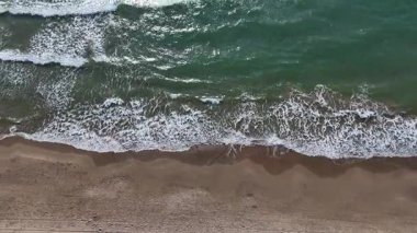 Yumuşak dalgalar kumlu bir plaja yuvarlanıyor köpüklü beyaz şapkaları öğle güneşinin altında parıldıyor. Sakin okyanus, mavi renkleri yansıtıyor. Sahilde huzur ve rahatlama hissi uyandırıyor..