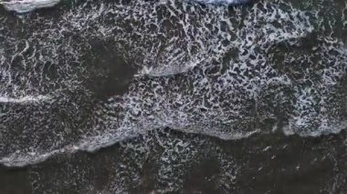 Dalgalar kıvrılır ve karanlık okyanus suyunda karmaşık desenler oluşturur. Güneşin yansımaları parıldıyor, öğleden sonra plaj atmosferinin huzur dolu güzelliğini arttırıyor..