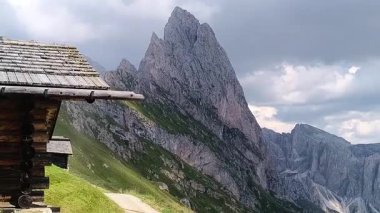 Kırsal bir ahşap kulübe ön planda duruyor, cazibesi yüksek bir dağ zirvesinin çarpıcı arka planıyla vurgulanıyor. Kara bulutlar gökyüzünde yuvarlanıyor, dingin manzaraya yoğunluk katıyor..