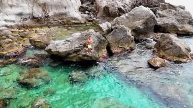 Bir insan, canlı deniz yaşamı ve engebeli kıyı manzarasıyla çevrili turkuaz suların ortasında, parlak güneş ışığı altında, saf mutluluk saçan büyük bir kayanın üzerinde güvenle durur..