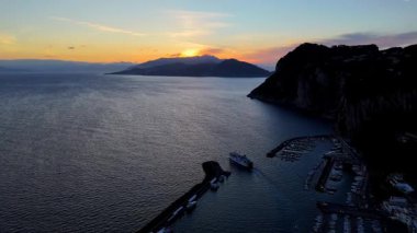 Güneş uzak tepelerin ardından batarken kıyıya doğru hafif dalgalar süzülür. Limanda kayıklar sallanıyor, gökyüzünü aydınlatan canlı renklerle. Huzurlu ortam, yansımayı ve rahatlamayı davet eder..