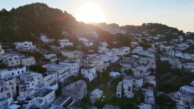 Güneş sakin bir yamaçtaki köyün üzerinden yükselirken, altın ışınları aşağıdaki tuhaf beyaz binaları ve canlı yeşil ağaçları aydınlatıyor ve kırsal alanda huzurlu bir sabah atmosferi yaratıyor..