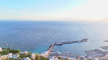 Sakin bir limana bakan kayıklar, güneş uzak dağların ardında yavaşça batarken suda sallanıyor, yakınlardaki beyaz yıkanmış binaların üzerine altın rengi bir parıltı saçıyor..