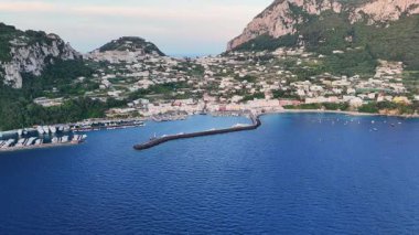 Overlooking the enchanting seaside town of Capri, the image captures colorful buildings nestled against lush green hills. Boats dot the harbor under a soft sunset glow, creating a serene atmosphere.