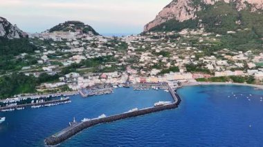 A stunning aerial view captures the lively marina of Capri, surrounded by lush greenery and charming buildings. The setting sun casts a warm glow over the tranquil waters, enhancing the beauty.