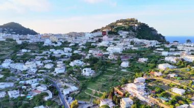 Nestled on the slopes of a stunning island, the hillside community showcases rows of white-washed homes and vibrant greenery, with the crystal blue sea in the background at dusk.
