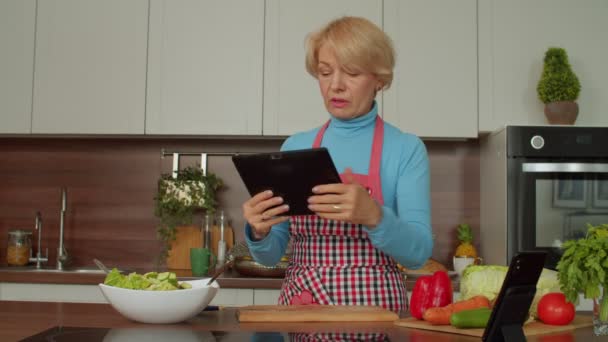 デジタルタブレットを使用してオンラインで夕食のレシピを閲覧エプロンで魅力的な正の引退した熟女 キッチンで健康的な食事を調理を楽しみながらおいしい新鮮な食材をチェック — ストック動画
