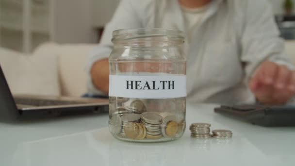 男性将硬币放入印有健康字样的玻璃瓶的特写镜头 而退休男性则管理财务和账单 并在室内为健康保险储蓄 — 图库视频影像