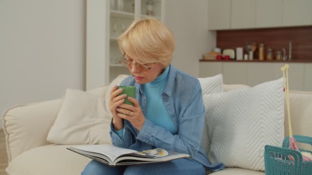 穿着眼镜坐在沙发上 喝着杯子里的热饮 翻阅着食谱 一边在自家房间里享受休闲 一边寻找食谱的迷人的退休成年女性 — 图库视频影像