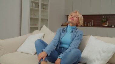 Huzurlu, emekli, günlük kıyafetleriyle yoga egzersizi yapan, kanepe üzerinde meditasyon yapan, stresi azaltmak ve enerjiyi yenilemek için iç odada dinlenen, son sınıf öğrencisi kadın..