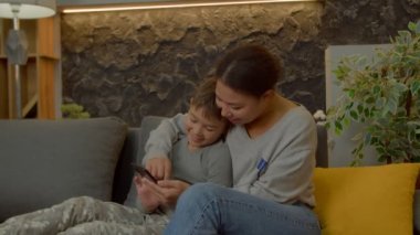 Olumlu, mutlu, çok kültürlü, ilköğretim yaşında bir çocuk ve pozitif, çekici siyah Asyalı bir anne kanepede dinleniyor, konuşuyor ve sosyal medya içeriğini internetten araştırıyor..