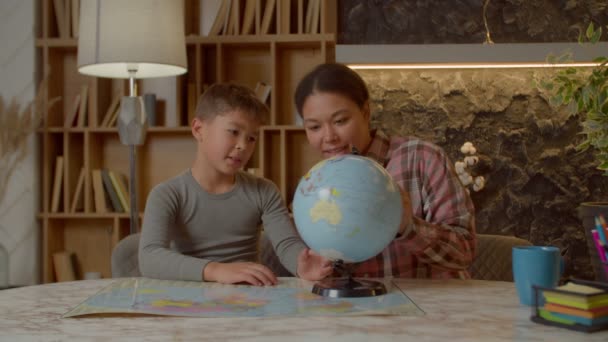 专心致志地投入聪明可爱的多民族学龄男孩 关心迷人的亚裔黑人母亲 一起学习地理 在全球地图上搜寻不同的国家 在家学习 — 图库视频影像