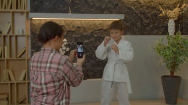 Sevimli, motive olmuş, karate kimonolu çoklu-ırk öncesi çocuk savaş duruşunda duruyor, kararlı bir ifadeyle bakarken gururlu siyah anne cep telefonuyla fotoğraf çekiyor..