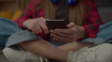 Orta kısımda oturan sevimli Latin kız kanepede bağdaş kurarak oturuyor, akıllı telefon kullanarak online oyun oynuyor ve evde boş vakitlerin tadını çıkarıyor. Yakın plan..