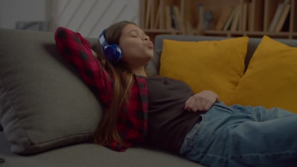 喜びのかわいいヒスパニック系の10代の女の子目はソファの上でリラックスしたワイヤレスヘッドフォンで閉じます 国内の部屋でレジャーを過ごしながら 歌をキャッチするために沿って音楽や歌を楽しみます — ストック動画