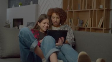 Neşeli, sevimli, çok kültürlü genç kız ve mutlu Afrikalı Amerikalı anne kanepede oturuyor, tablet bilgisayarını paylaşıyor, iletişim kuruyor ve sosyal medya içeriğini içeride dinlenirken tartışıyorlar..