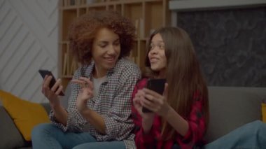Afro-Amerikan pozitif çekici anne ve neşeli, çoklu etnik çeşitliliğe sahip genç kız cep telefonlarıyla iletişim kuruyor, internette geziniyor ve evde dinlenirken sosyal medya içeriğini tartışıyor..