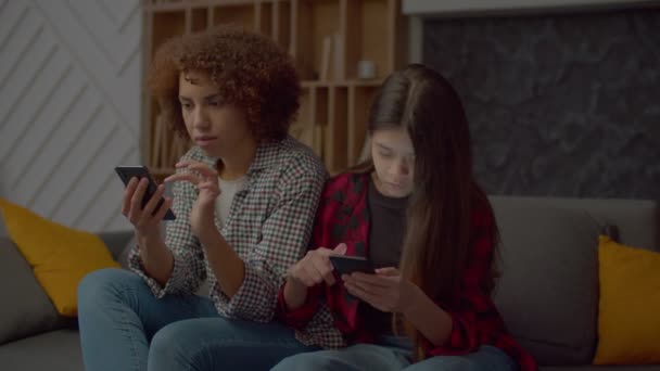 スマートフォンの愛らしい多人種の10代の娘と魅力的な黒の母親に隣接し 国内の部屋でソファに座っている間 ネットワーキングとオンラインでコンテンツを閲覧 — ストック動画