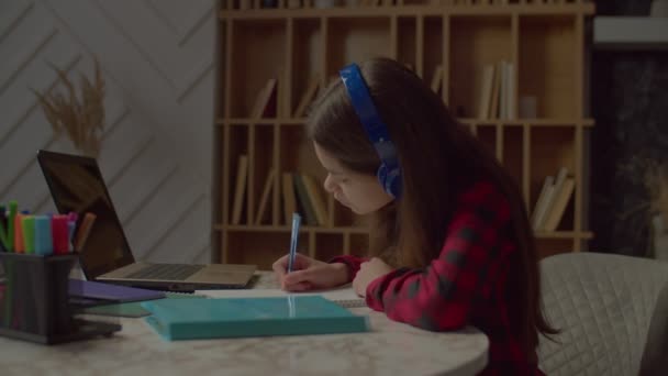 在无线耳机中学习的聪明可爱的拉丁少女用笔记本电脑远程在线学习课程 在家学习时在练习本上写作业的侧视图 — 图库视频影像