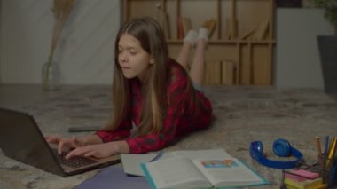 Tatlı zeki İspanyol ergen kız dizüstü bilgisayar ve ders kitabı ile çalışıyor, ortaokul için ödev hazırlıyor ve egzersiz kitabına ödevlerini yazarken yerde uzanıyor..