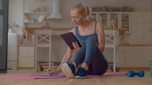 漂亮迷人的老年女性适合坐在运动垫上 与数字平板电脑联网 计划健身锻炼 同时准备在室内进行体育锻炼 — 图库视频影像