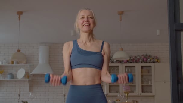 積極的にやる気のある魅力的なスポーティなフィットシニア女性は 国内の部屋で体重トレーニングを練習しながら 上腕の筋肉を強化し ダンベルBicepカール運動を行う — ストック動画