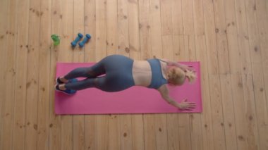 Aktif motivasyonlu çekici spor fitnesinin üst görüntüsü olgun kadın aşağı doğru köpek tekmesi egzersizi yapıyor, alt vücudu esnetiyor ve kapalı alanda egzersiz yaparken üst gövdeyi güçlendiriyor..
